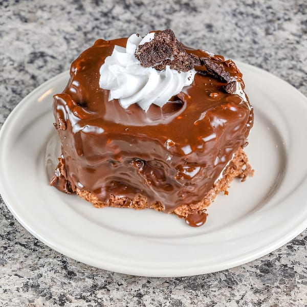 A rich chocolate cake with thick chocolate icing, whipped cream, and brownie crumbles on a white plate.