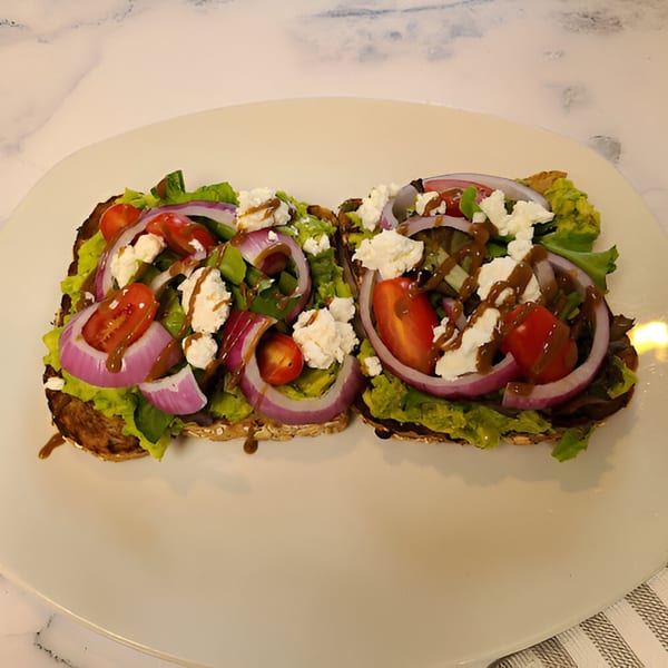 Two avocado toasts with red onion, cherry tomatoes, feta, and balsamic glaze on a white plate.