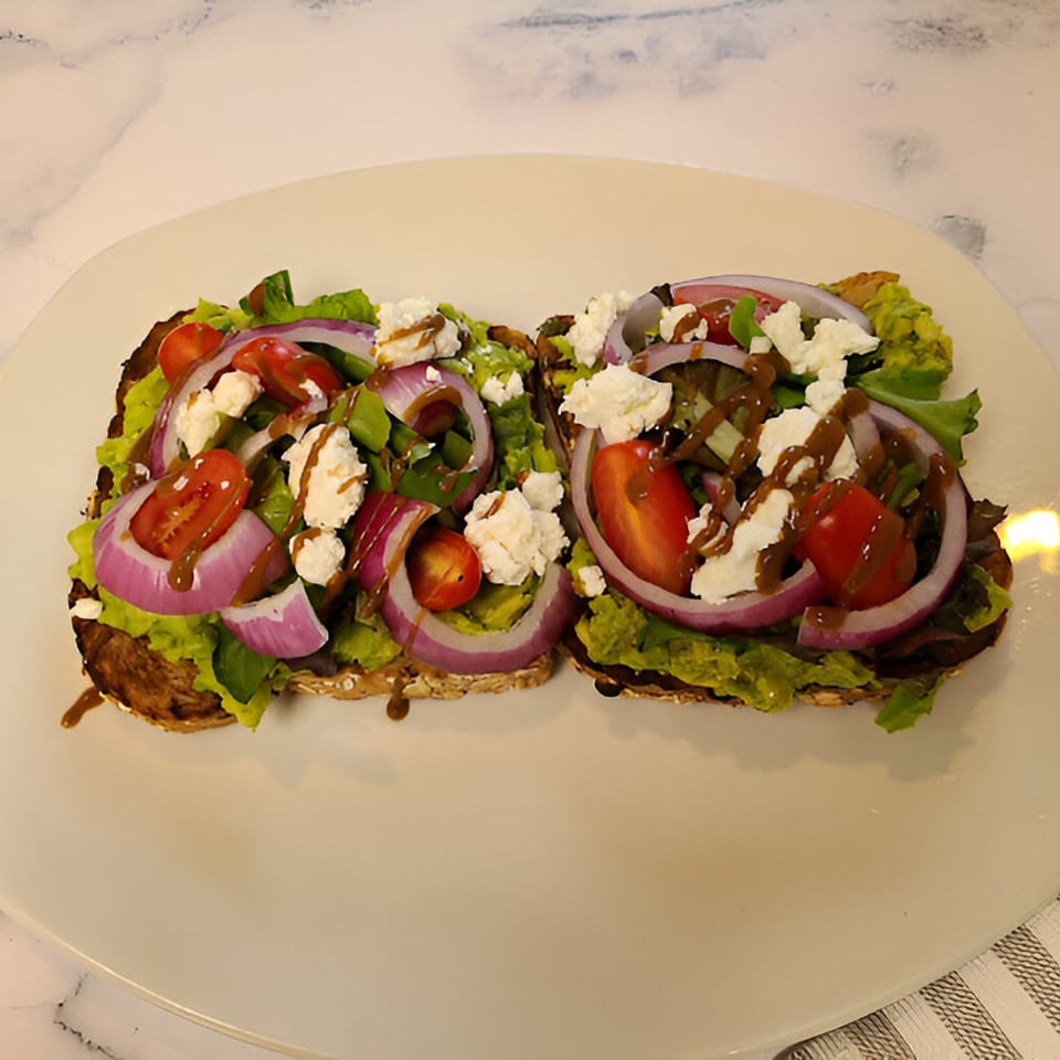 Two avocado toasts with red onion, cherry tomatoes, feta, and balsamic glaze on a white plate.