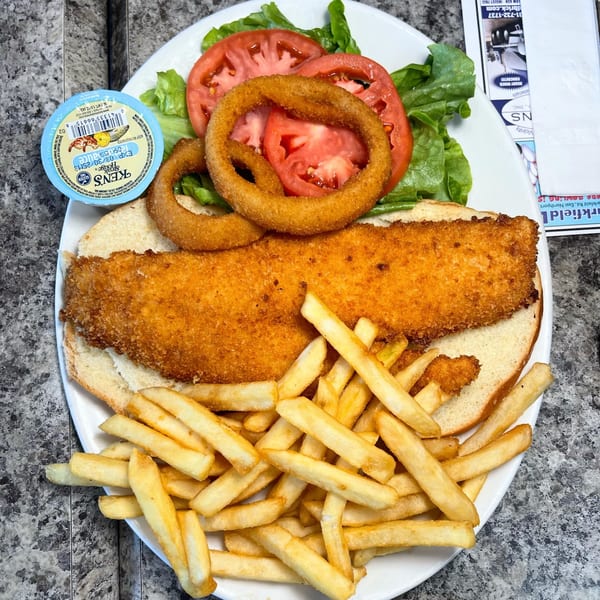 A fried fish sandwich with lettuce, tomato, onion rings, fries, and tartar sauce on a white plate.