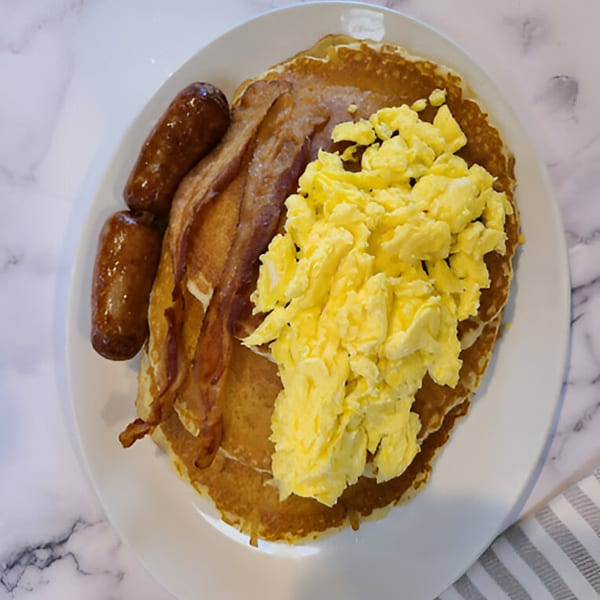 A diner breakfast plate with pancakes, scrambled eggs, bacon, and two sausage links.