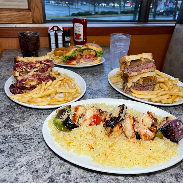Diner table with plates of pastrami sandwich, cheeseburger, shrimp skewers, and fries.